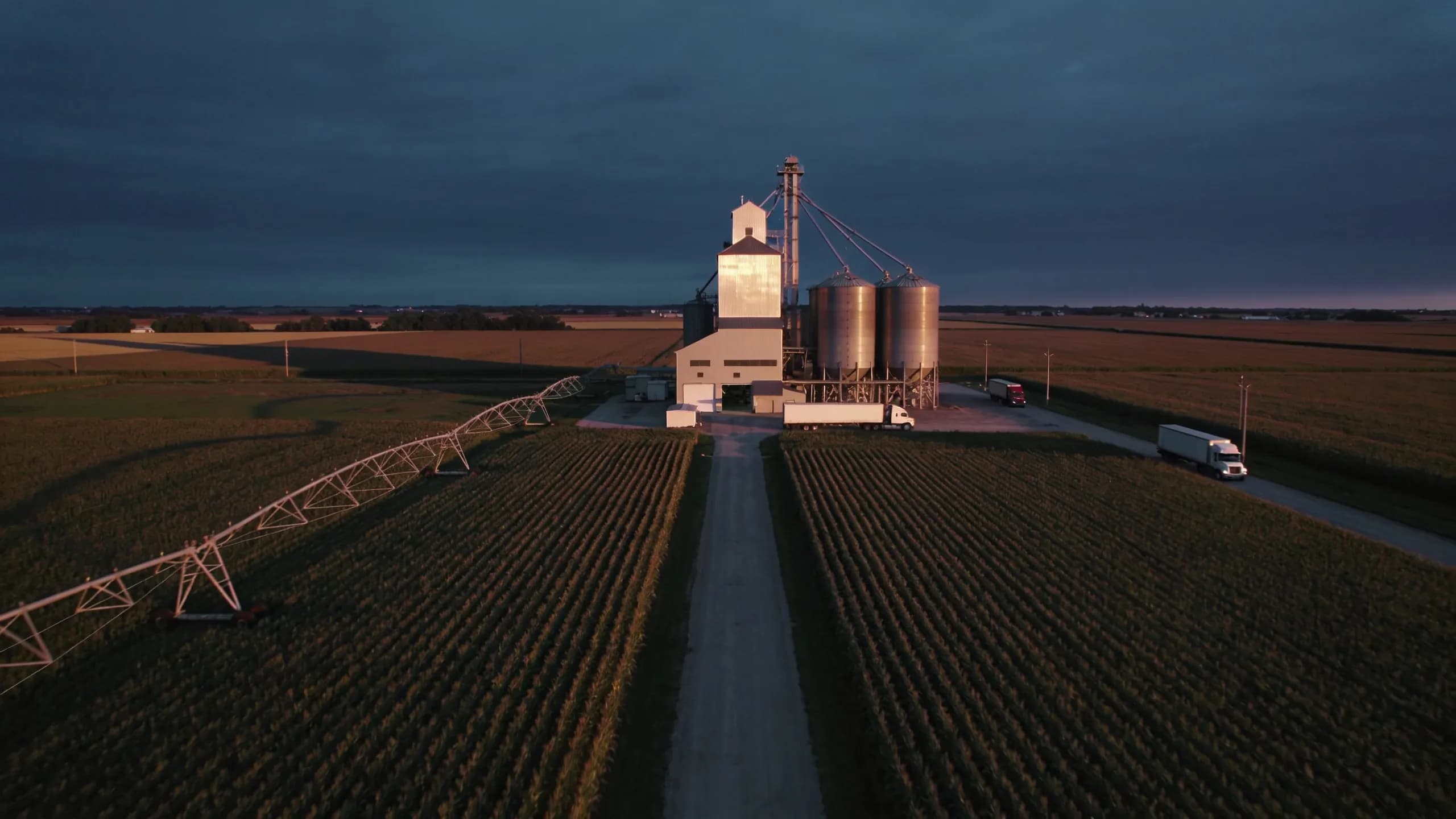 Aerial view of a commercial agricultural operation at dusk, crop rows converging toward a processing facility